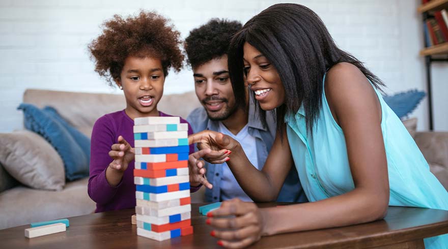 Een gezin speelt een torenspel met houten blokken op een tafel. De blokken zijn rood, blauw en wit.