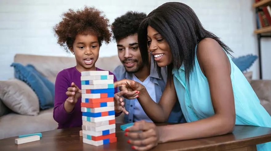 Een gezin speelt een torenspel met houten blokken op een tafel. De blokken zijn rood, blauw en wit.