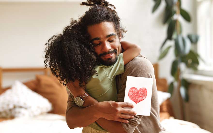 A tender scene of a man hugging a child, smiling softly while holding a card decorated with a bright red heart.