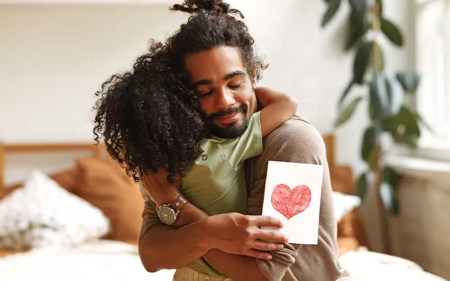 A tender scene of a man hugging a child, smiling softly while holding a card decorated with a bright red heart.