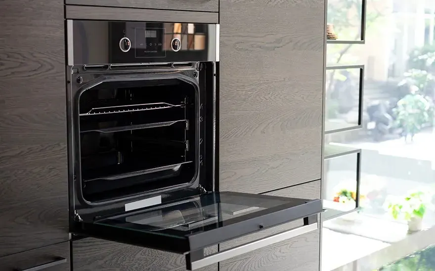 Open inbouwoven met glazen deur omlaag, donkere houten keukenkast en veel daglicht van grote ramen naast het moderne toestel.