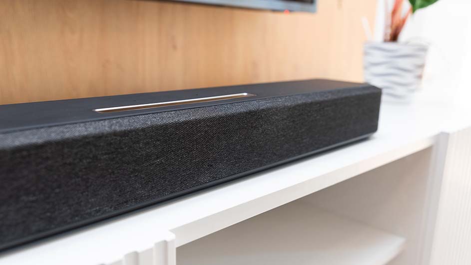 Zwarte soundbar met een zilveren lijn erop, staat op een witte kast tegen een houten achtergrond met een plant.