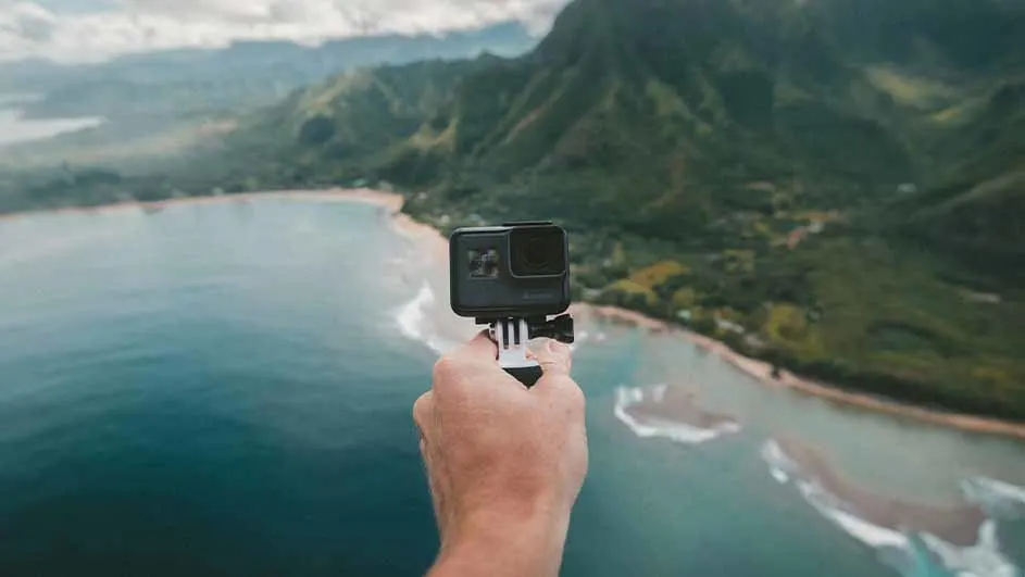 Een hand houdt een camera vast met een kustlijn en groene bergen op de achtergrond.