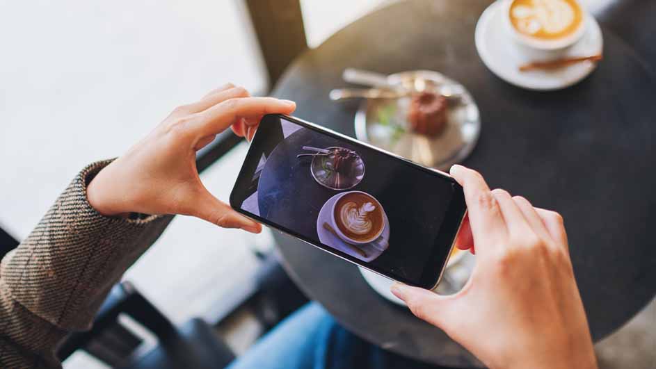 Persoon fotografeert koffie en gebak op een zwarte tafel met een smartphone. Een tweede kop koffie staat op de achtergrond.