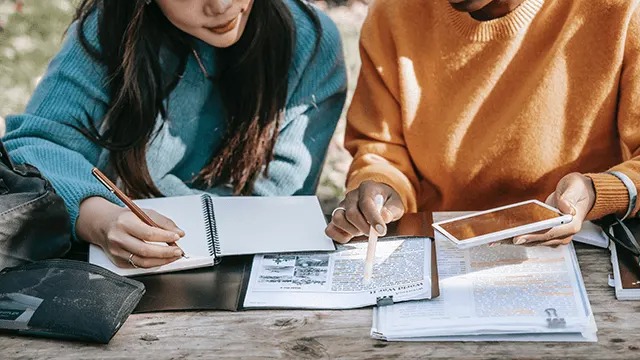 Two people study outdoors, writing in notebooks with a Samsung phone on the table, highlighting text in printed documents and sharing ideas.