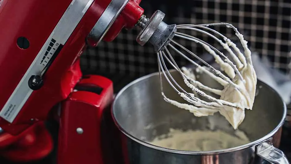 Een rode keukenmachine met een metalen kom en een garde vol beslag. De achtergrond heeft een geruit patroon.