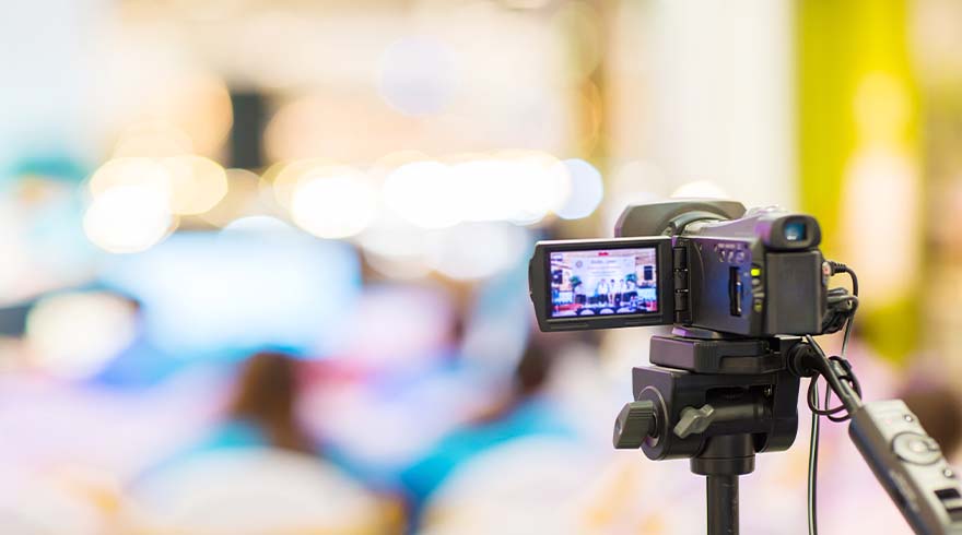 Een zwarte videocamera op een statief, met een scherm dat een scène weergeeft, tegen een vage achtergrond.