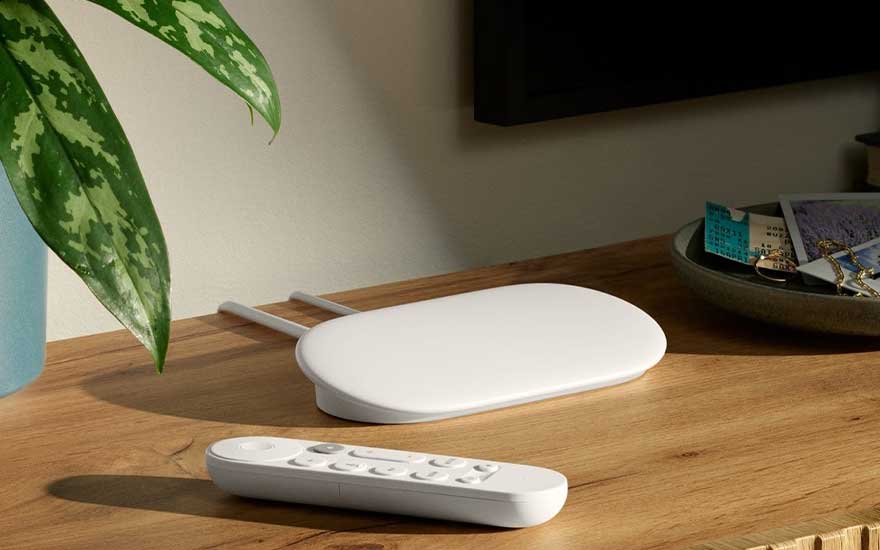 White streaming media player with remote control on wooden table beside plant and decorative bowl.