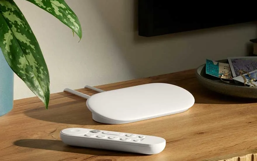 White streaming media player with remote control on wooden table beside plant and decorative bowl.