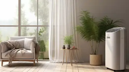 A beige armchair with a blanket is near a window. There's a table, plants, and a portable air conditioner.
