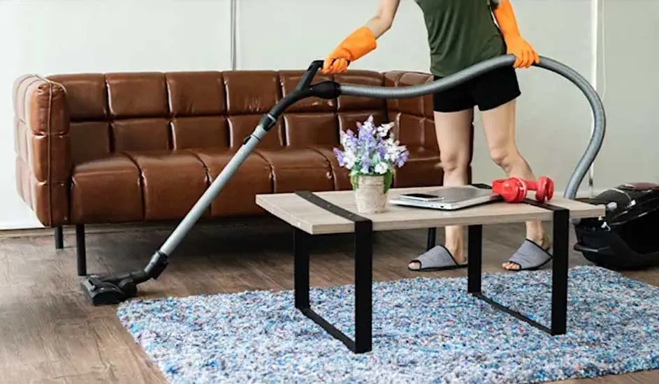 Iemand stofzuigt een blauw vloerkleed voor een bruine leren sofa, met oranje handschoenen aan en een tafel vol laptop, bloemen en gewichten.