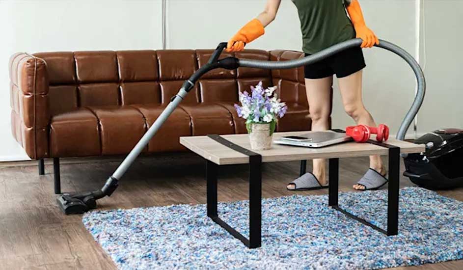 A person vacuums a blue rug by a brown leather sofa, wearing orange gloves, beside a coffee table with laptop, flowers and dumbbells.