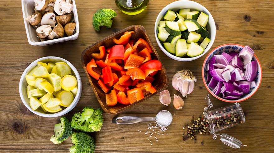 Diverse groenten, zoals paprika, courgette, ui, broccoli en champignons in schalen op een houten tafel, met kruiden.