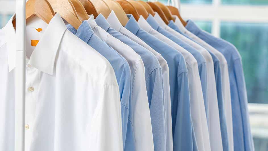 Een rij witte en blauwe overhemden hangt aan houten hangers aan een metalen rek, voor een wazige achtergrond.