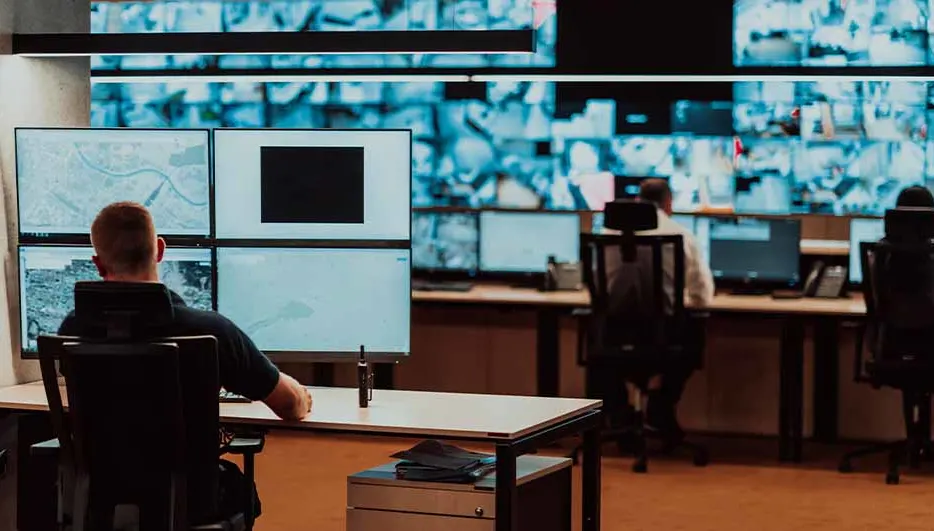 Een controlekamer met mensen achter bureaus die naar meerdere computerschermen kijken.