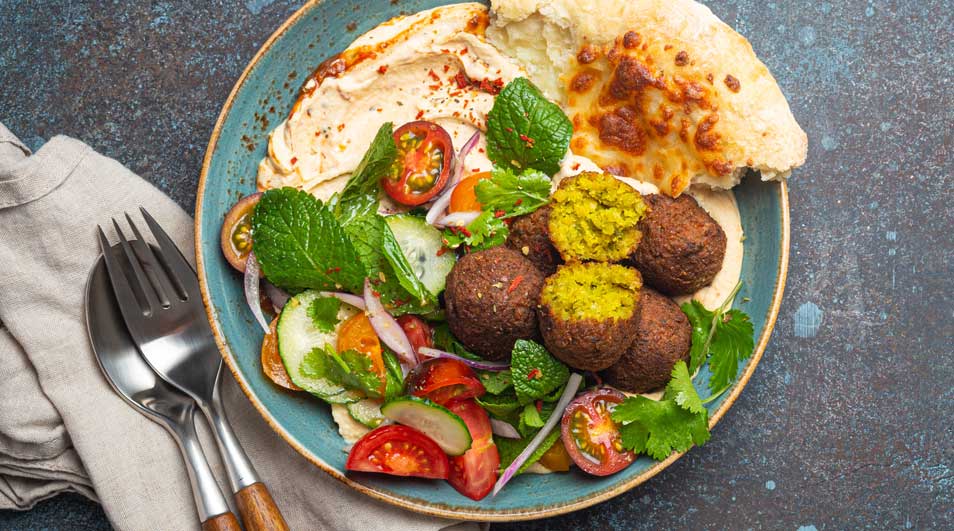 Een blauw bord met falafel, pita, hummus, komkommer, tomaat en kruiden. Een vork en lepel liggen naast een beige servet.