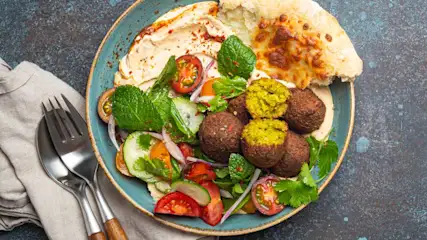 Een blauw bord met falafel, pita, hummus, komkommer, tomaat en kruiden. Een vork en lepel liggen naast een beige servet.