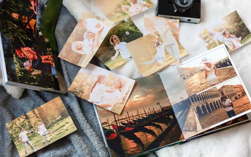 Photos and albums are scattered on a gray blanket. Images include landscapes, family moments, and travel scenes. A black camera is visible.