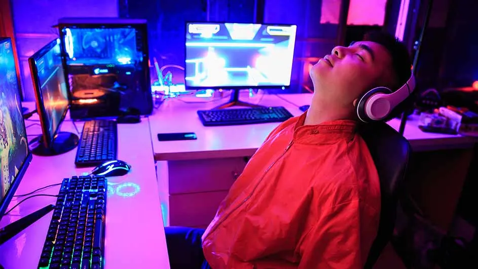 Een persoon met een rode jas en een koptelefoon zit achter een bureau met computers en toetsenborden.