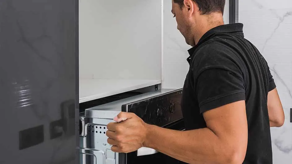 Een man in zwart shirt installeert een zilverkleurige oven in een keukenkast.