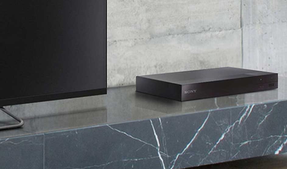 Op een grijze marmeren kast staat een zwarte Sony mediaspeler naast een tv, tegen een betonnen muur.