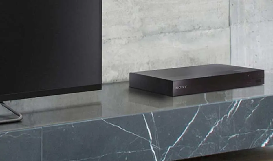 Op een grijze marmeren kast staat een zwarte Sony mediaspeler naast een tv, tegen een betonnen muur.