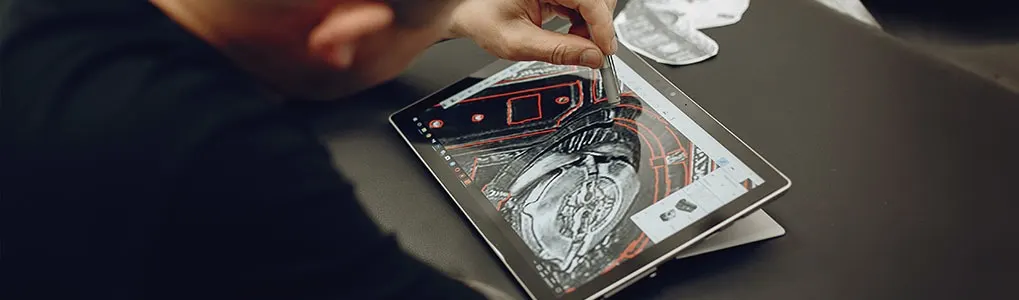 A person uses a stylus on a tablet showing a detailed drawing, with a dark surface in the background.
