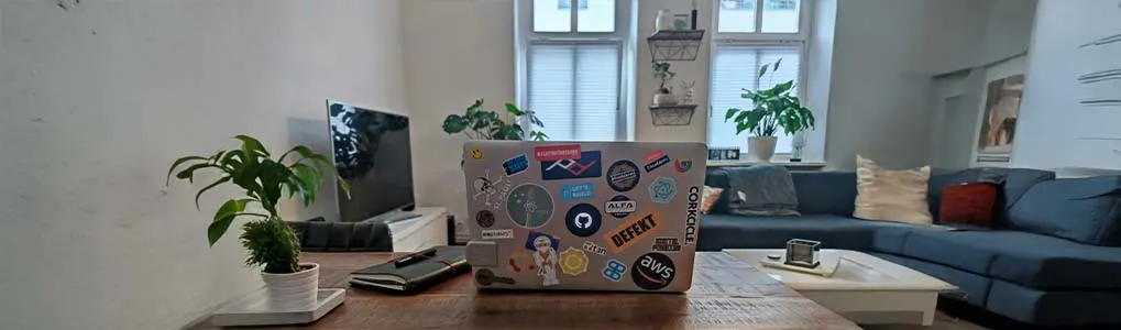 Een MacBook met allerlei stickers op de achterkant staat op een houten tafel in een woonkamer met veel planten, een bank en een televisie op de achtergrond.