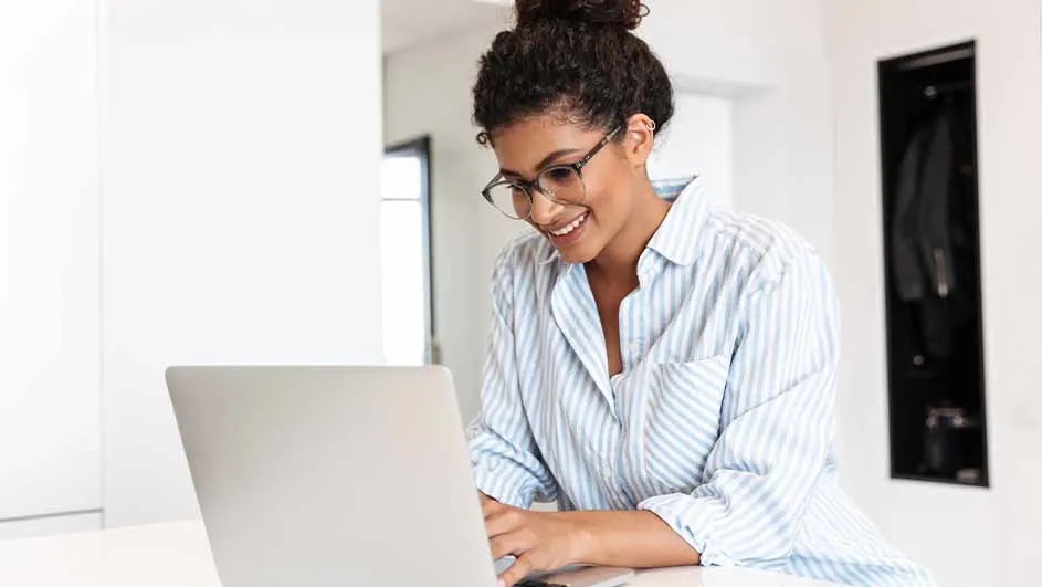 Vrouw met bril en gestreept overhemd, lachend achter een laptop. Op de achtergrond een witte muur en een zwarte kast.