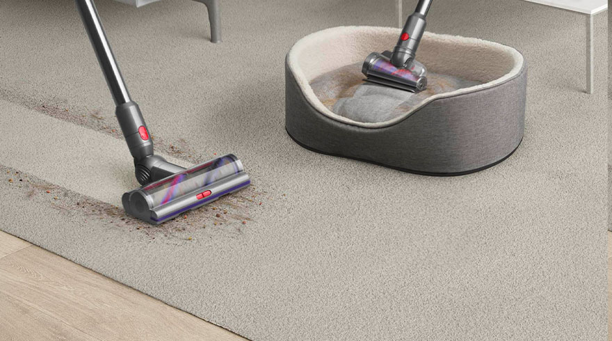 Een grijze stofzuiger reinigt een beige tapijt en een grijze hondenmand met witte rand.