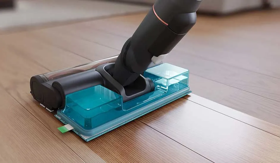 A mop attachment on a cordless stick vacuum glides across the wooden floor, using a blue water tank to wet clean the surface.
