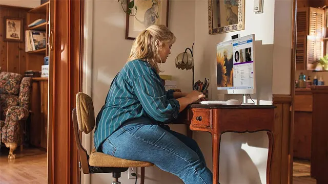 Vrouw zit aan een houten bureau voor een computer. Naast haar staat een stoel en een houten kast.