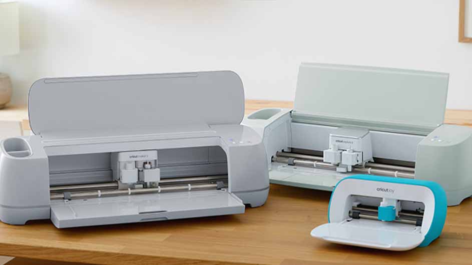 Drie Cricut-machines staan op een houten tafel. De machines zijn wit en blauw en hebben open kleppen.