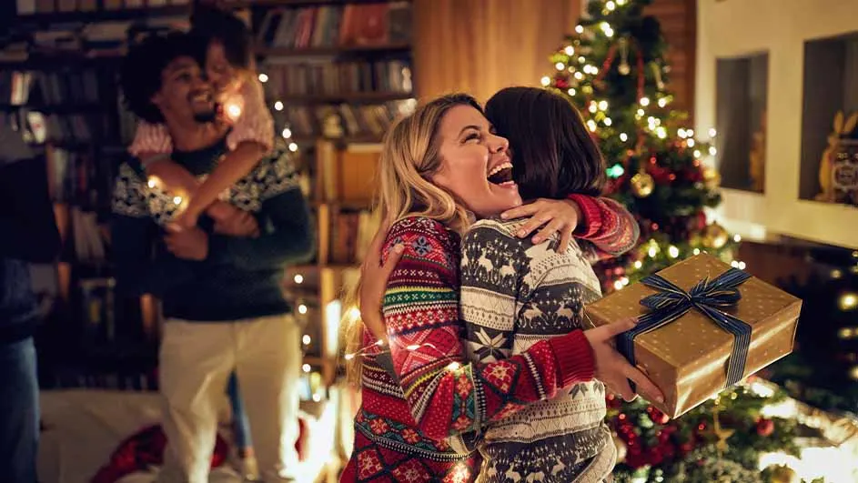 Twee vrouwen omarmen elkaar bij een kerstboom, één houdt een cadeau vast. Een man draagt een kind op zijn rug op de achtergrond.