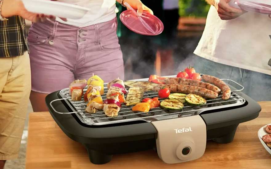 People gather around an electric Tefal grill with sausages, skewers and grilled vegetables, ready to share a cosy meal.