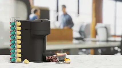 Een zwarte koffiemachine staat naast groene en gele capsules. Een kopje koffie en bonen staan op een witte tafel.