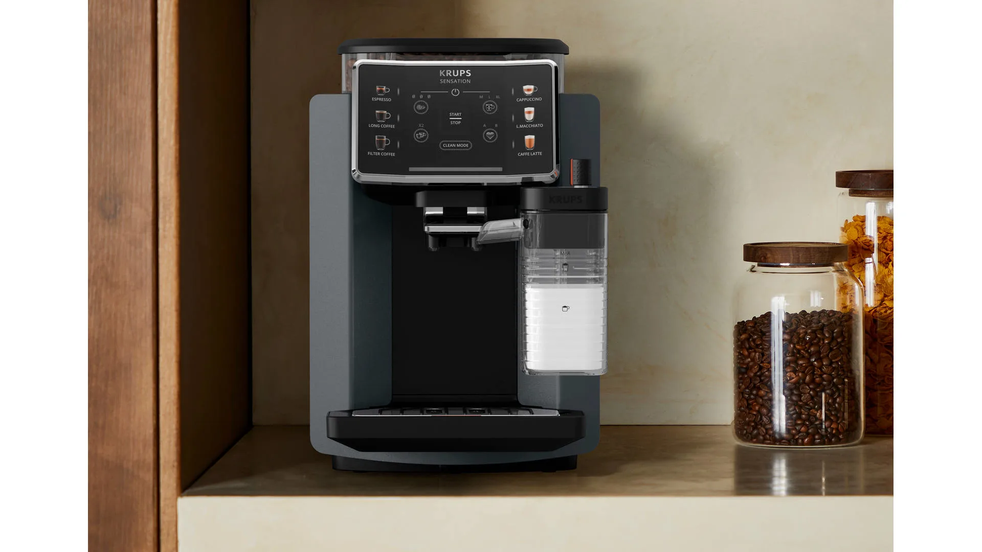 Grijze Krups koffiemachine met melkreservoir. Op de achtergrond staan glazen potten met koffiebonen en ontbijtgranen.