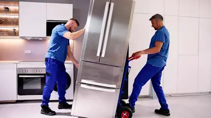 Two workers in blue uniforms move a stainless steel refrigerator with a dolly in a white kitchen.