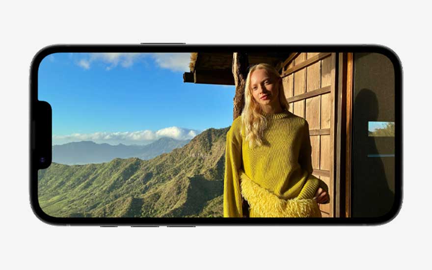 Een iPhone in liggende stand toont een foto op volledig scherm van een vrouw in een gele trui, poserend voor een houten hut in berglandschap.