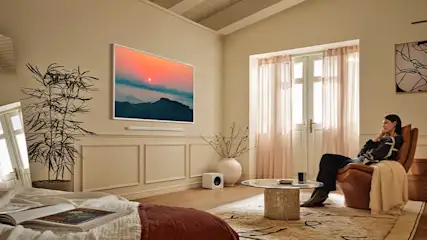 Een vrouw zit ontspannen op een stoel in een lichte kamer. Aan de muur hangt een tv die een kleurrijke zonsondergang boven bergen toont.