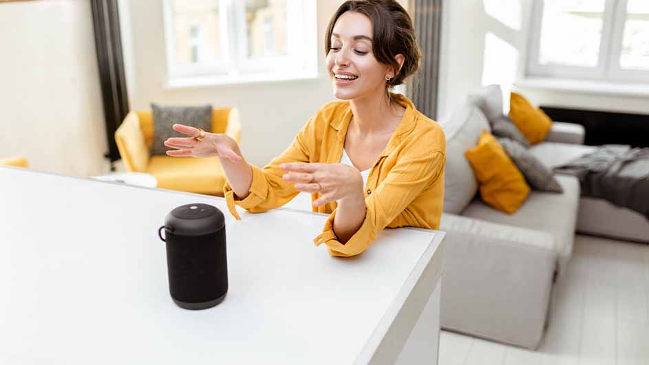 Vrouw in gele blouse gebaart naar een zwarte speaker op een wit oppervlak. Achtergrond: gele stoel en grijze bank.