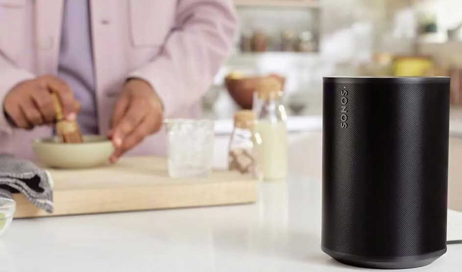 On a white kitchen counter stands a black Sonos speaker while someone prepares a dish with fresh ingredients in the background.