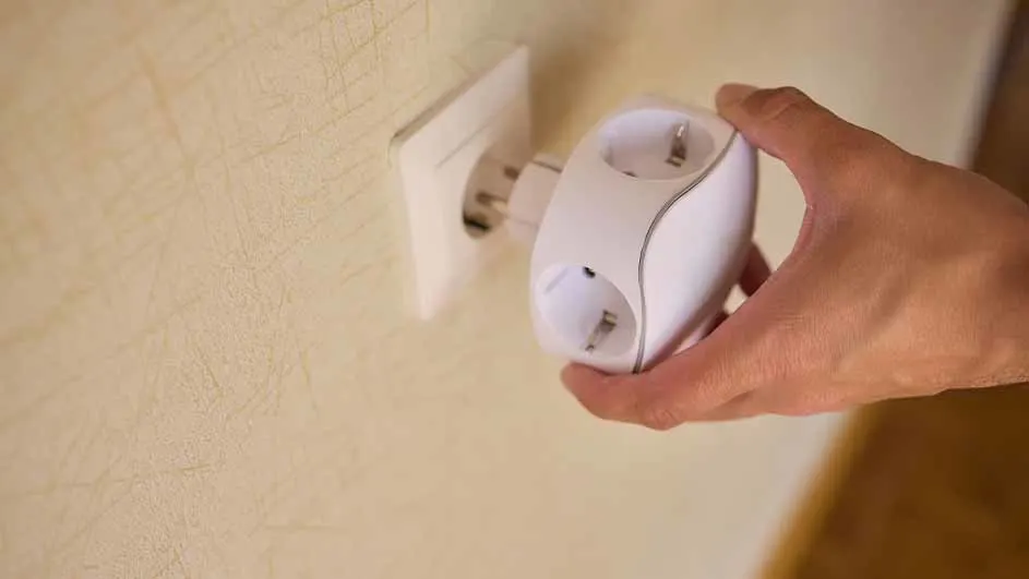 Hand steekt wit stopcontact met drie aansluitingen in een ander stopcontact tegen een beige muur.
