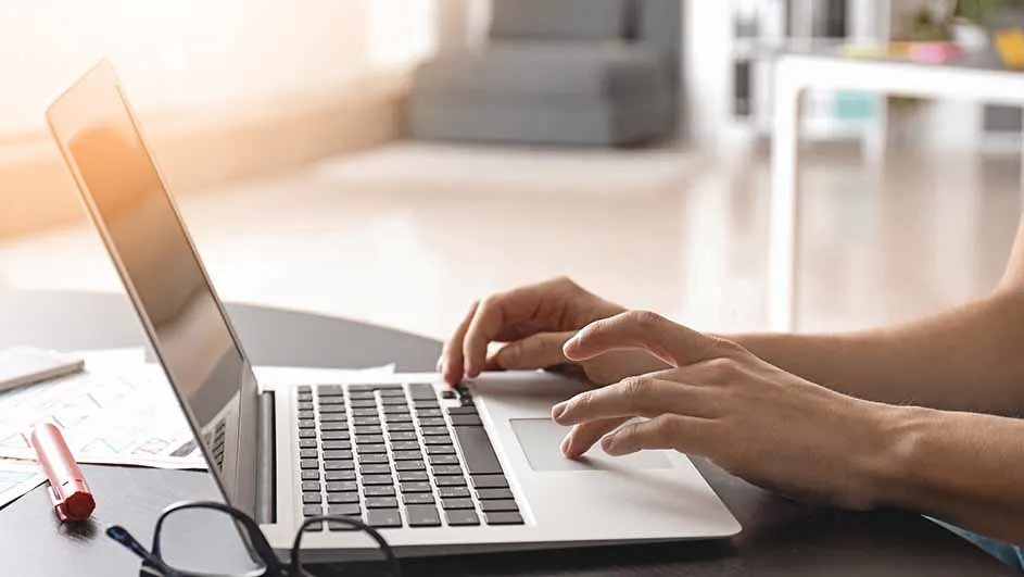 Een persoon typt op een laptop, met een bril, een pen en documenten op het bureau. De achtergrond is licht en vaag.