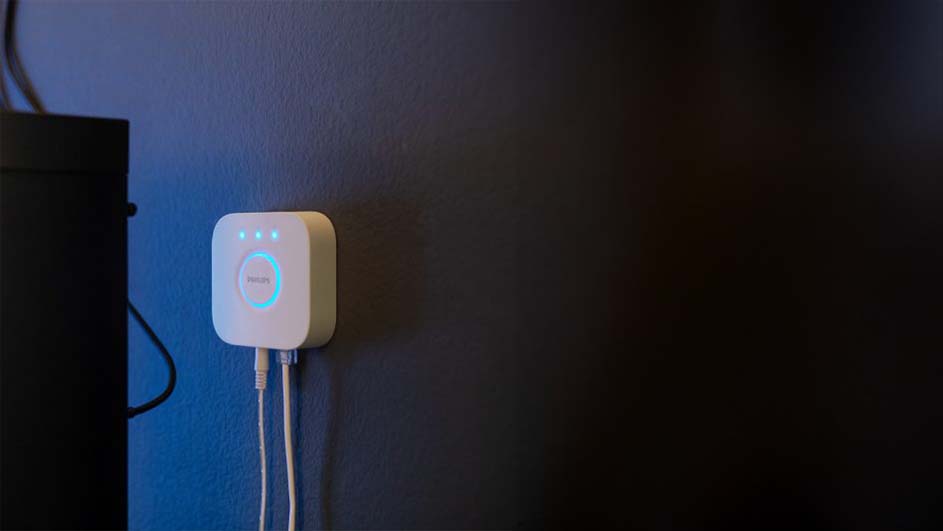 Witte Philips Hue-bridge aan een blauwe muur met twee witte kabels eronder, en blauwe lichtjes aan de voorkant. Zwarte rand links.