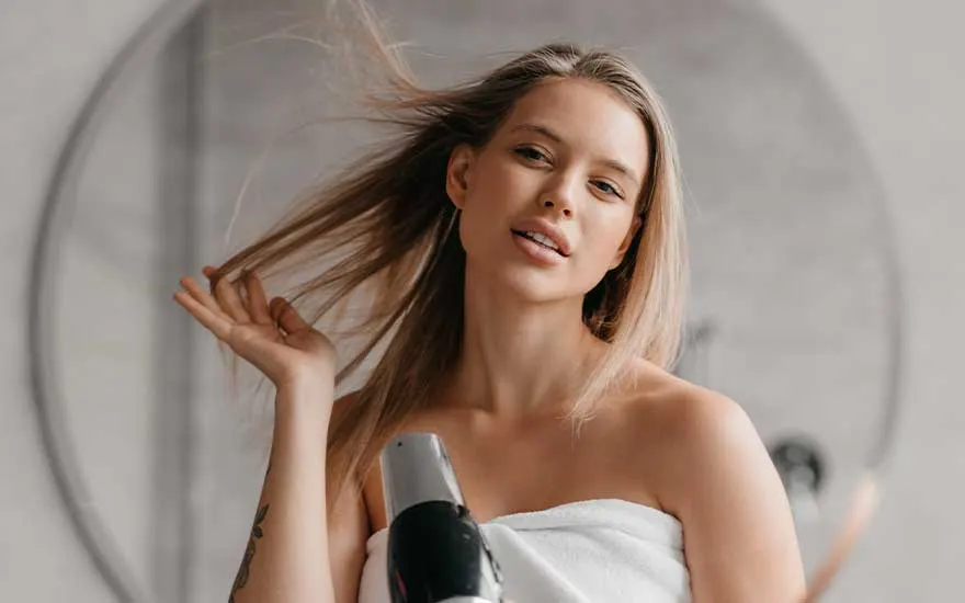 Een jonge vrouw in een witte handdoek droogt haar lange haar met een föhn en kijkt zelfverzekerd in de spiegel van de badkamer.