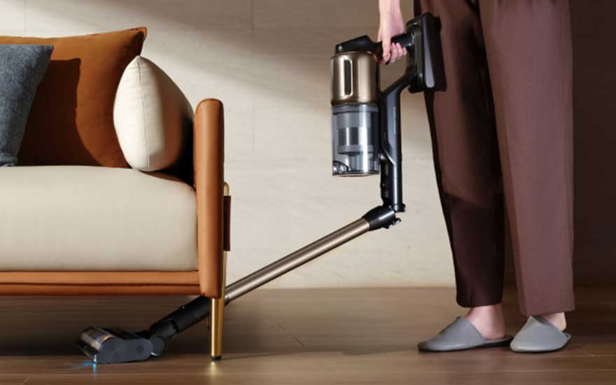 Iemand gebruikt een flexibele steelstofzuiger om onder een sofa te reinigen, dicht bij de poot en houten vloer.