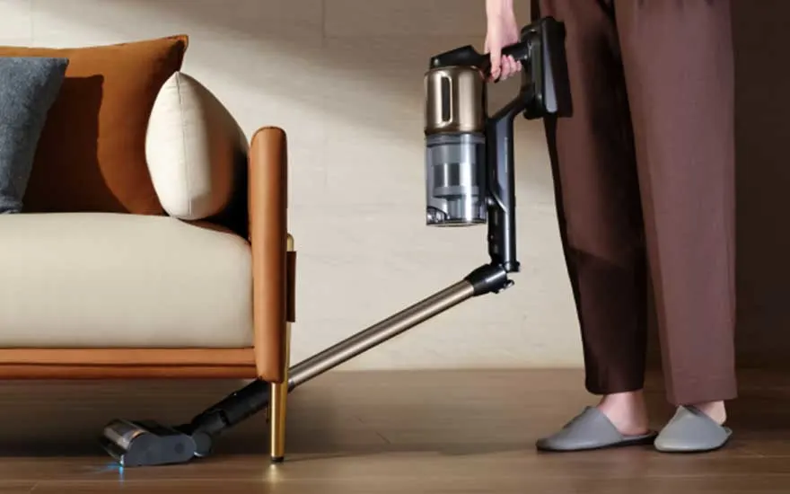 Iemand gebruikt een flexibele steelstofzuiger om onder een sofa te reinigen, dicht bij de poot en houten vloer.