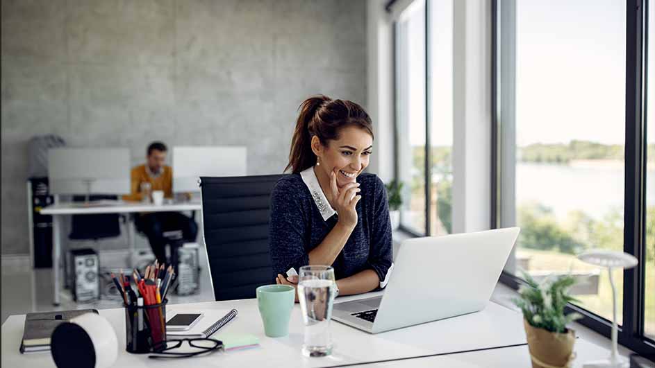 Vrouw lacht naar laptop op bureau met beker, waterglas en plant. Man zit aan bureau op achtergrond, naast groot raam.