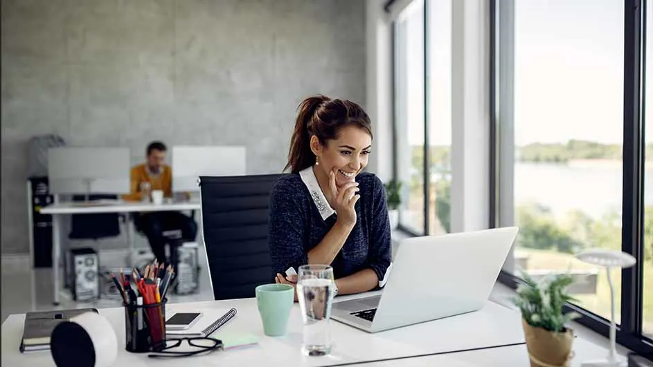 Vrouw lacht naar laptop op bureau met beker, waterglas en plant. Man zit aan bureau op achtergrond, naast groot raam.